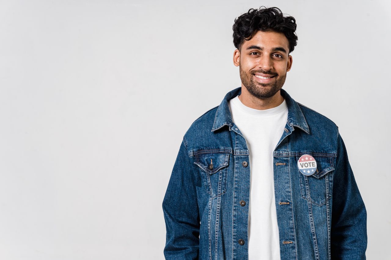 A smiling man wearing a denim jacket with a vote button on a white background.