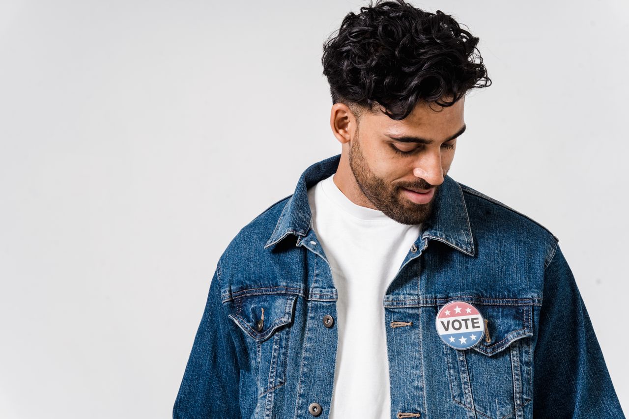 Man with a vote button on his denim jacket, looking down.